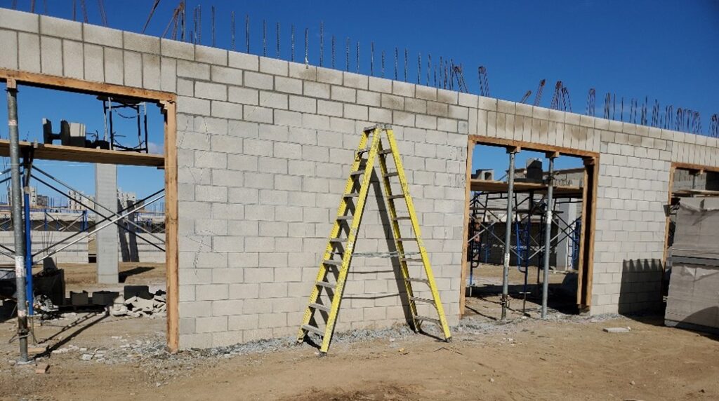 Construction site featuring a partially built wall with a yellow ladder leaned against it, surrounded by scaffolding and construction materials.