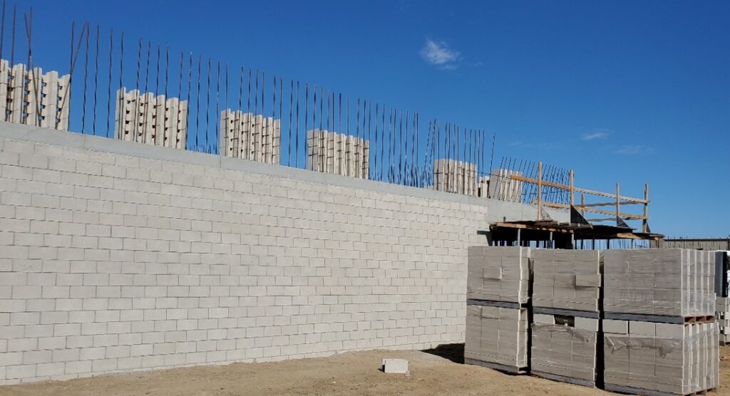 Construction site showing a concrete block wall with rebar protruding from the top, stacks of blocks nearby, and a scaffold in the background.